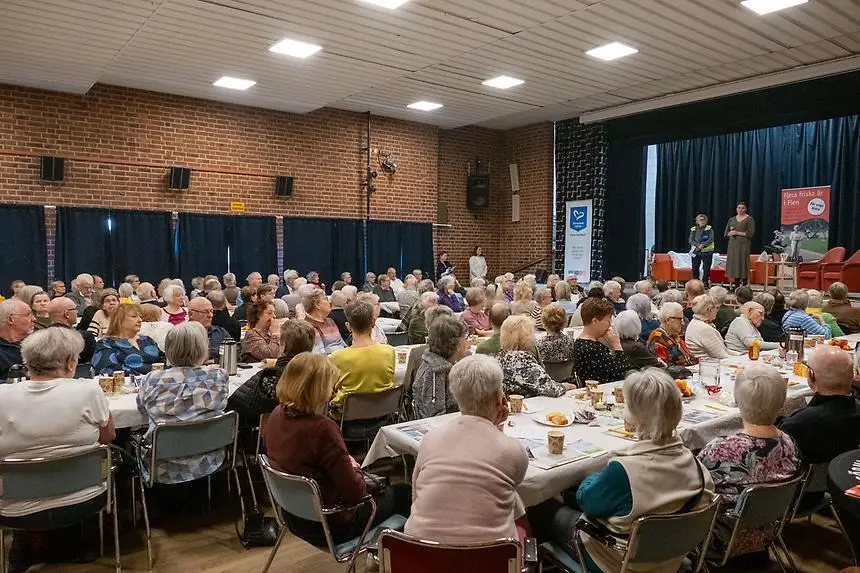 En samling personer sitter runt bord med mat i en hall.