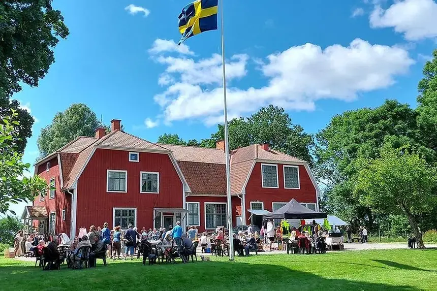 Ett rött trähus med ett blått och gult flagg på en hög flaggstång. Människor samlas på gräsmattan framför huset under en blå himmel.