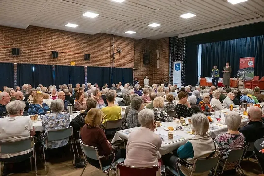 En samling personer sitter runt bord med mat i en hall.
